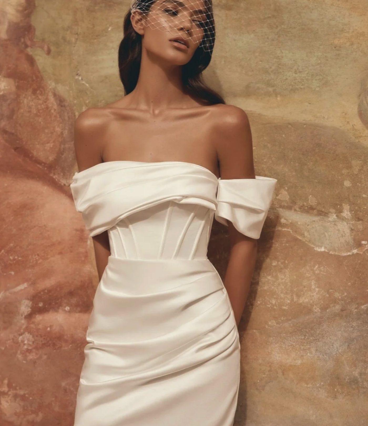 A bride in an off-the-shoulder structured mini dress against a warm stone wall