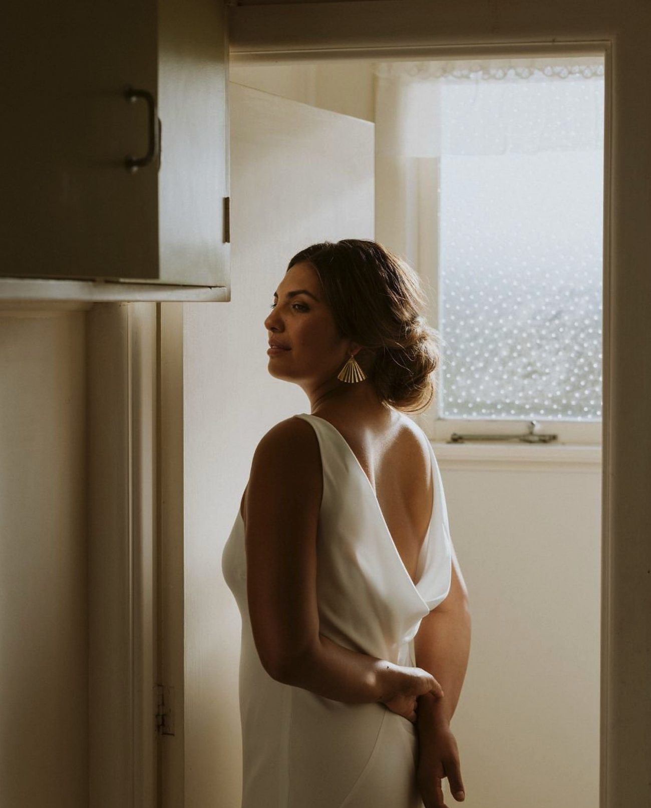 A bride in a minimal draped gown standing by a window