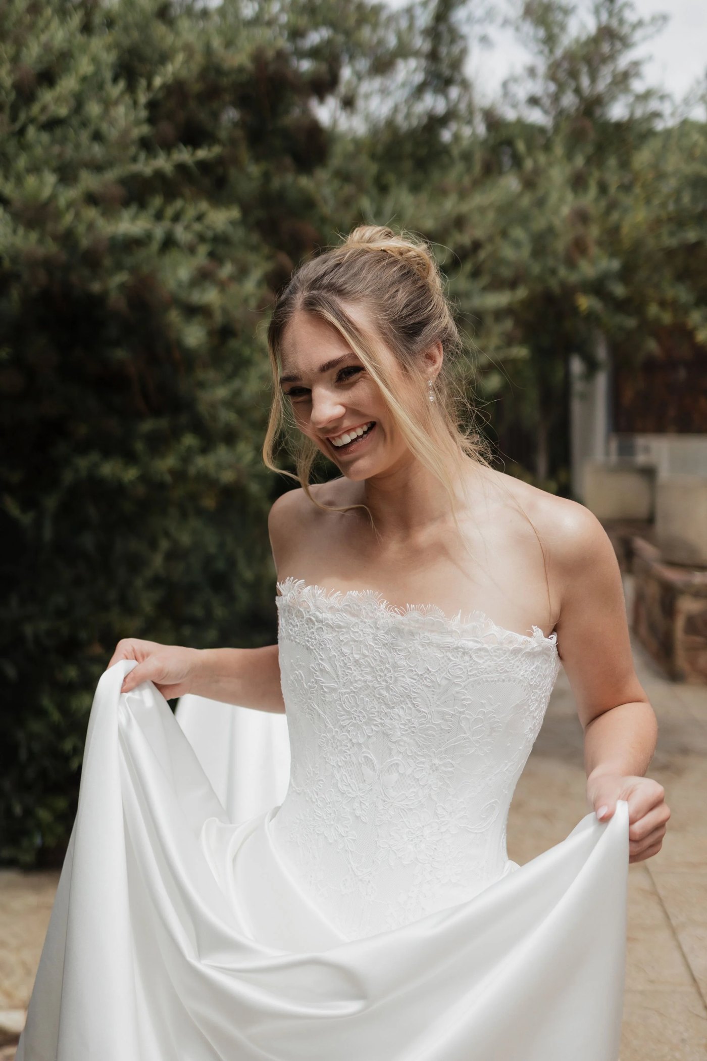 A laughing bride lifting the skirt of a strapless lace and satin gown by Elli Nicole